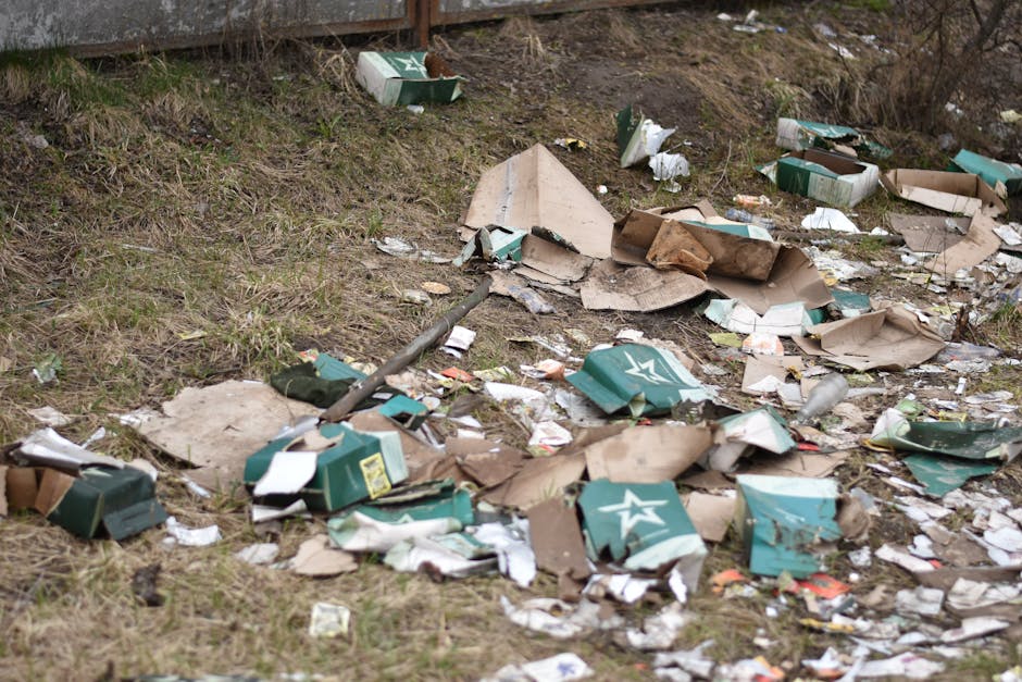 A person wearing a turquoise disposable glove is seen picking up a clear plastic bottle from a grassy outdoor area. In the background, a yellow and white plastic bag filled with waste is visible, slightly blurred. The person, whose arm and part of their leg are visible, appears to be engaged in litter collection, emphasizing a community effort to clear outdoor rubbish. The grass beneath is green with some small patches of dirt, and the scene is lit naturally, indicating daytime. The environment suggests a park, garden, or roadside verge where waste is being gathered for disposal. This image aligns with services offered by Rubbish Removal Catford, which focuses on independent waste collection and on-site clearance to help maintain clean and tidy outdoor spaces, supporting environmentally responsible rubbish removal solutions in local areas.