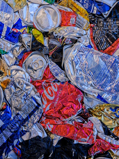 A close-up view of a large pile of crushed aluminum and plastic beverage cans, primarily silver, with some red, blue, yellow, and black labels visible. The cans display various brand markings, with one prominent Coca-Cola can in the centre showing the red logo. The cans are tightly compressed and stacked irregularly, with some partially folded or crushed, and others showing pull tabs or flat tops. The surface textures include smooth, shiny metal and matte paper labels, reflecting ambient light. The background shows additional cans and discarded packaging, suggesting an environment of waste collection or recycling preparation, potentially on a pavement or disposal site. The scene exemplifies waste materials ready for secondary processing, illustrating the importance of proper rubbish removal and disposal services in private waste handling or alternative rubbish collection approaches. Rubbish Removal Catford's service might involve the collection and sorting of such recyclable discarded cans, supporting local waste management efforts while providing an environmentally responsible method of waste disposal.