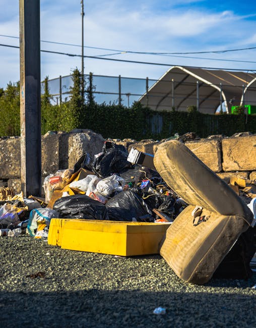 A large pile of mixed domestic waste, including black and white plastic rubbish bags, discarded cardboard and paper, and a worn-out, beige car seat, all strewn across a gravel surface in an outdoor area. The waste is partially enclosed by a low stone wall made of irregularly shaped, weathered stones, with some debris spilling onto the ground. In the background, a tall metal pole is visible alongside overhead wires, and a hedge provides a natural backdrop. Further behind, a contemporary greenhouses or sports facility structure with a curved roof can be seen, suggesting an urban or semi-urban environment. The scene is lit by daylight, with a partly cloudy sky overhead, indicating daytime. The image conveys an impression of uncollected refuse in an area that could be serviced by private waste removal specialists such as Rubbish Removal Catford, highlighting the need for professional rubbish clearance to prevent clutter and maintain clean, accessible outdoor spaces.