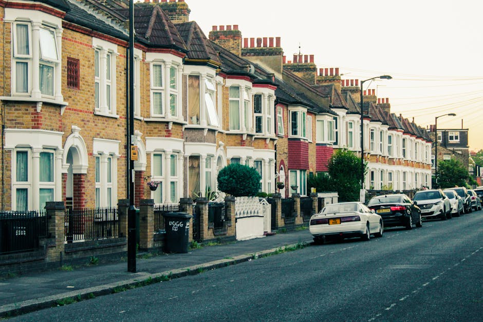 A row of Victorian terraced houses on Brownhill Road with brick facades in shades of yellow, red, and brown, each featuring white-framed bay windows and decorative architectural details. The houses have small front gardens enclosed by low brick walls topped with black metal railings, with some gardens containing neatly trimmed shrubs and potted plants. A few houses show open windows or minor exterior details such as hanging baskets. Along the pavement, several parked cars in various states of cleanliness and condition line the street, including sedans and hatchbacks in colors like white, black, and silver. A black wheelie bin marked ‘DOGG’ stands near one fence, indicating private waste collection by a local rubbish removal service. The scene is illuminated by soft daylight, with a clear sky and visible overhead power lines stretching into the distance, suggesting a quiet residential area where independent waste handling and rubbish removal are common alternatives to council services. This setting aligns with practical rubbish disposal efforts typical of property owners managing waste outside of local authority collection schedules, supporting services such as those provided by Rubbish Removal Catford.