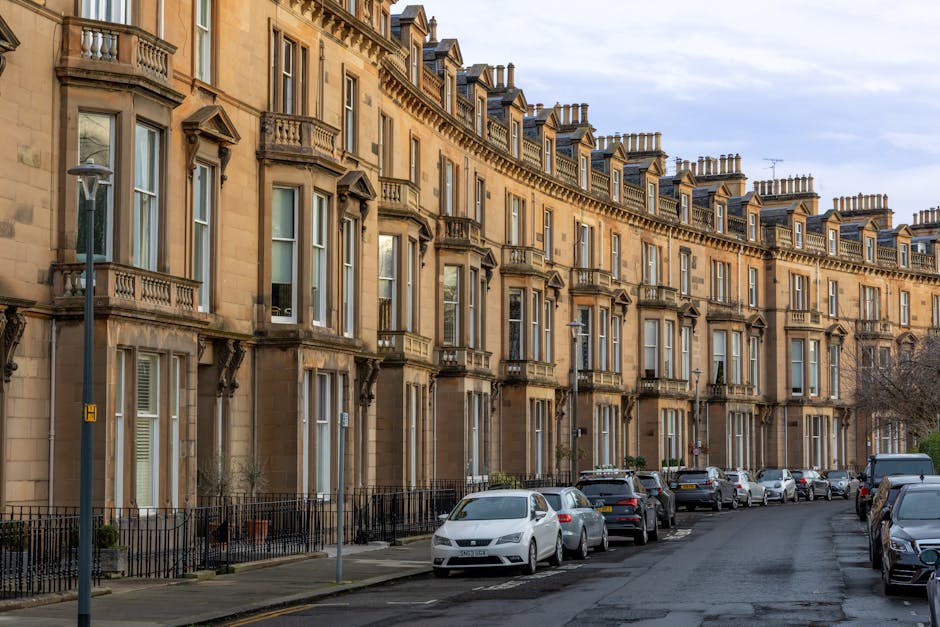 A row of three to four-storey Victorian terrace buildings constructed from light brown sandstone with ornate stone window surrounds, decorative cornices, and small balconies with wrought iron railings. The buildings have tall, narrow sash windows and pitched slate roofs with numerous chimney stacks. In front of the buildings, there is a paved sidewalk lined with black wrought iron railings, separating the footpath from the street. A series of parked cars, including sedans and hatchbacks in various dark and light colours, are aligned along the street, which appears to be a residential area. A large street lamp with a blue H sign is positioned on the pavement, and a single leafless tree is visible near the street corner, suggesting winter or early spring. The overall scene is illuminated by natural daylight, with a partly cloudy sky overhead, typical of urban residential zones where independent waste collection or private rubbish removal services like those offered by Rubbish Removal Catford may operate to manage refuse from these Victorian properties, ensuring cleanliness and compliance with local waste disposal standards.