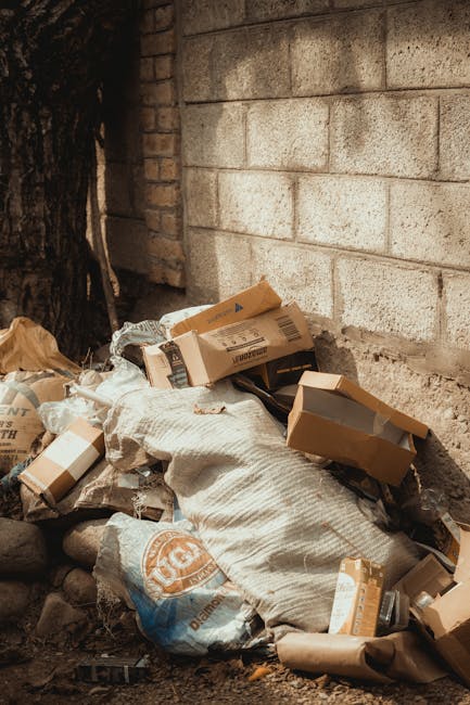 A pile of various discarded packaging materials and waste, including cardboard boxes, paper bags, and plastic wrapping, is situated at the base of a rough, grey concrete brick wall. Some boxes are partially collapsed or torn, with their flaps open or bent. A large white bag made of woven fabric lies amidst the debris, appearing slightly crumpled. The waste is resting on uneven ground composed of soil and small rocks, with a dark, weathered tree trunk visible on the left side of the image, indicating an outdoor setting. The wall behind the rubbish is constructed from concrete blocks with a coarse texture and light grey coloration, contrasting with the darker, organic materials of the waste. The scene is lit by natural daylight, casting soft shadows on the debris, highlighting the different textures and materials, and subtly conveying the concept of independent waste collection or local rubbish removal, similar to services offered by Rubbish Removal Catford, in a neutral, factual manner.