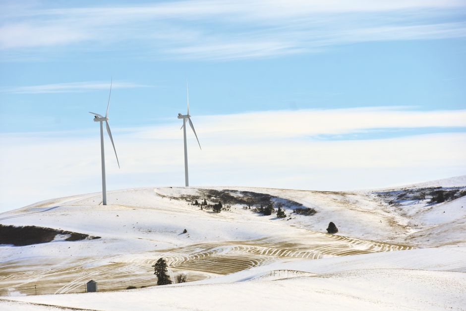 A winter landscape featuring two white wind turbines with three blades each, positioned on snow-covered rolling hills under a partly cloudy sky with shades of blue and white clouds. The terrain displays patches of dark forest and exposed earth contrasting with the snow, with visible lines from agricultural activity or landscape features across the foreground. Sparse trees are scattered along the slopes, and the scene is illuminated by natural daylight, creating a clear and calm atmosphere. This setting illustrates rural countryside with renewable energy infrastructure, relevant for observations on private waste handling or on-site management scenarios in environmentally conscious regions, as reflected in discussions about alternative waste disposal options. The image captures the vast openness of the landscape, emphasizing both the natural environment and man-made structures, naturally aligning with themes of off-grid or independent energy and waste solutions in the context of rubbish removal services in the UK.
